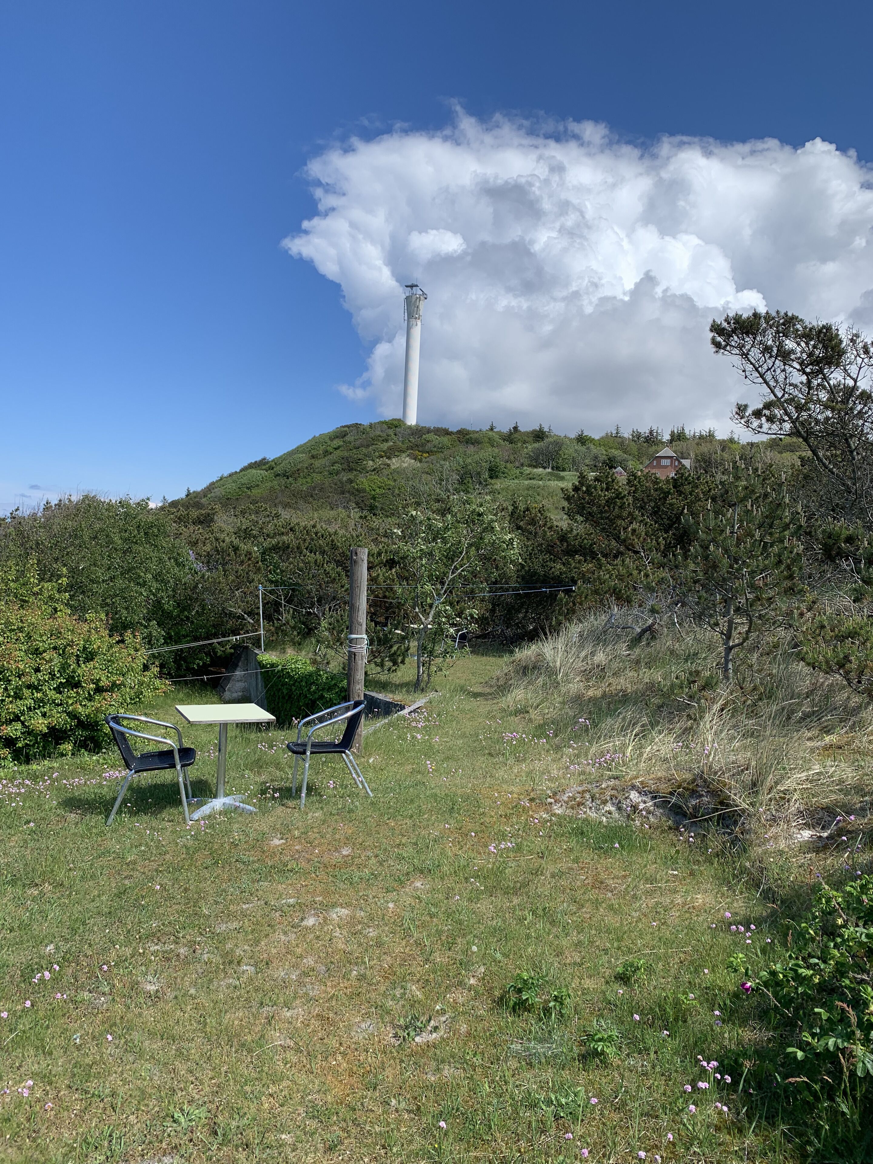 A grassy area with a metal table and chairs, a tall wind turbine in the background, and a house on a hill.