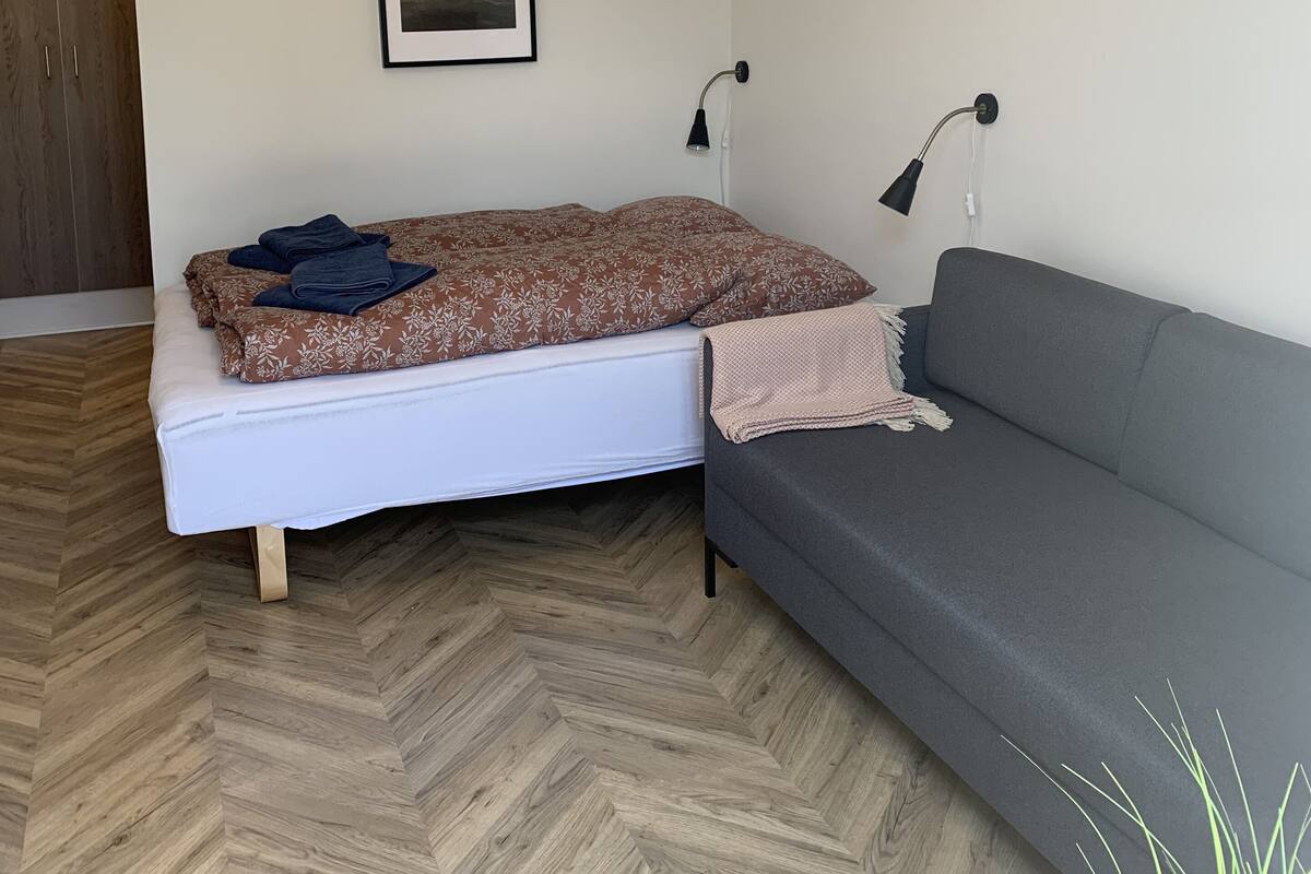 Une pièce comprenant un lit, un canapé gris, une armoire en bois, deux lampes fixées au mur et deux tableaux encadrés.