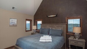 Un dormitorio con una cama grande, dos mesitas de noche, una mesita auxiliar, un cuadro enmarcado y una ventana con persianas.