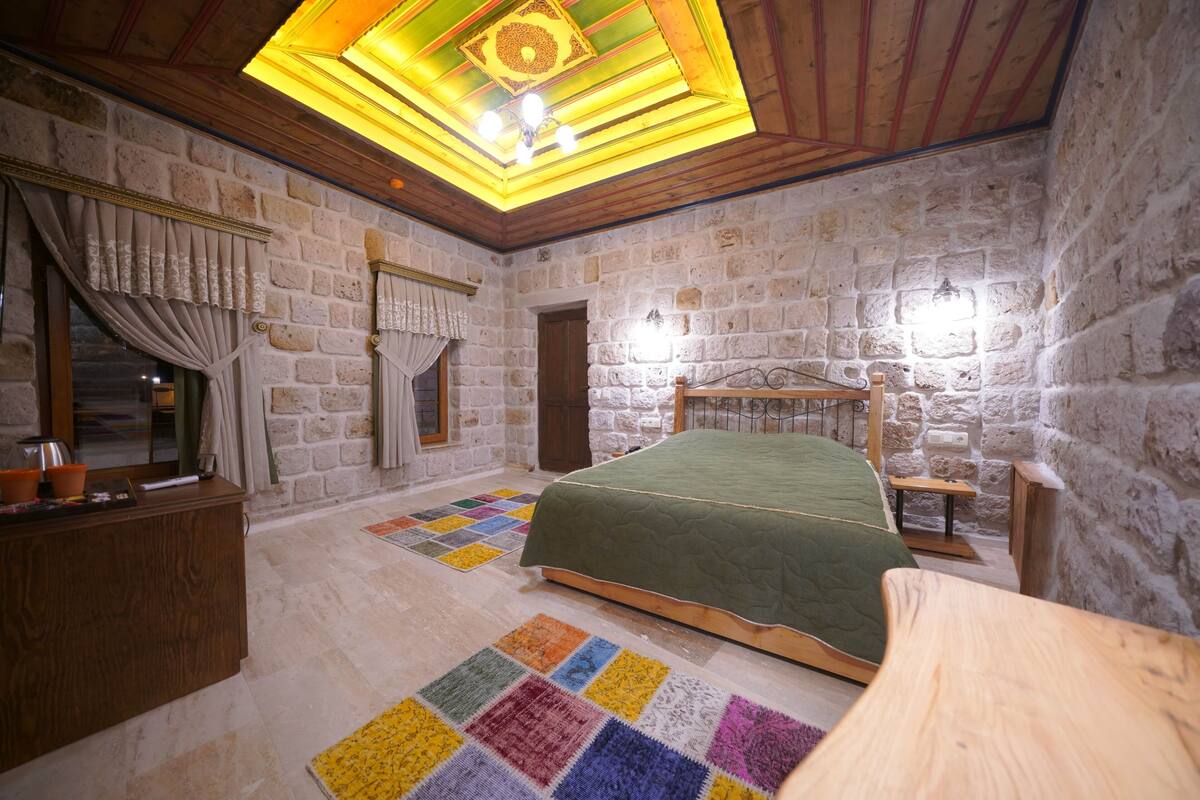 A bedroom with a stone wall, wooden ceiling, a bed with a green bedspread, a wooden dresser, and a colorful rug.