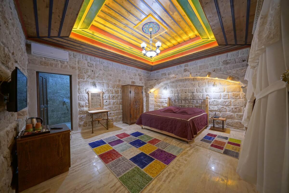 A room with a wooden ceiling, stone walls, a bed with a red bedspread, a wooden nightstand, a television, and a colorful rug.
