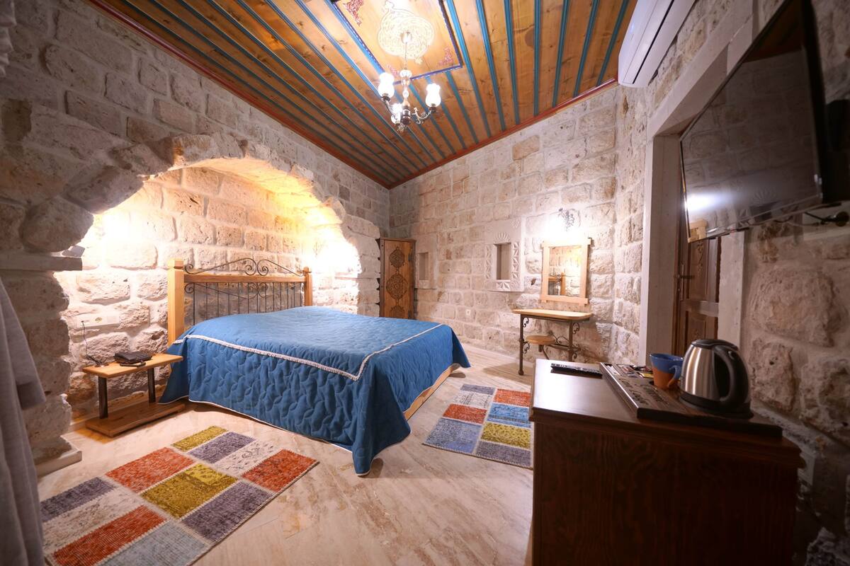 A stone-walled bedroom with a wooden ceiling, a bed with a blue quilt, a small table with a kettle, and a colorful rug.