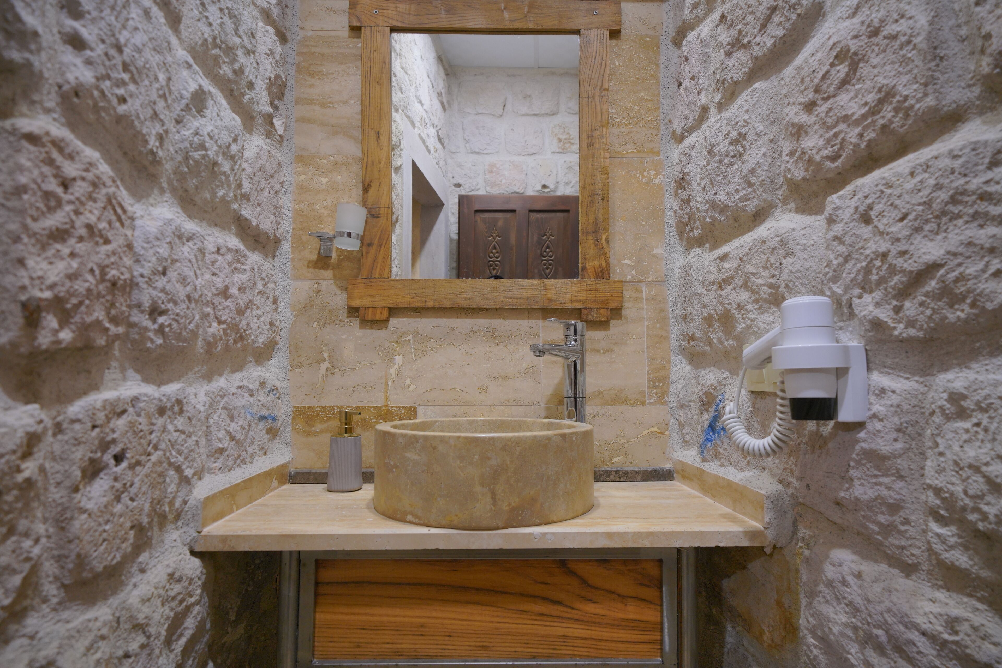 Un baño con pared de piedra, espejo con marco de madera, lavabo redondo y secador de manos.