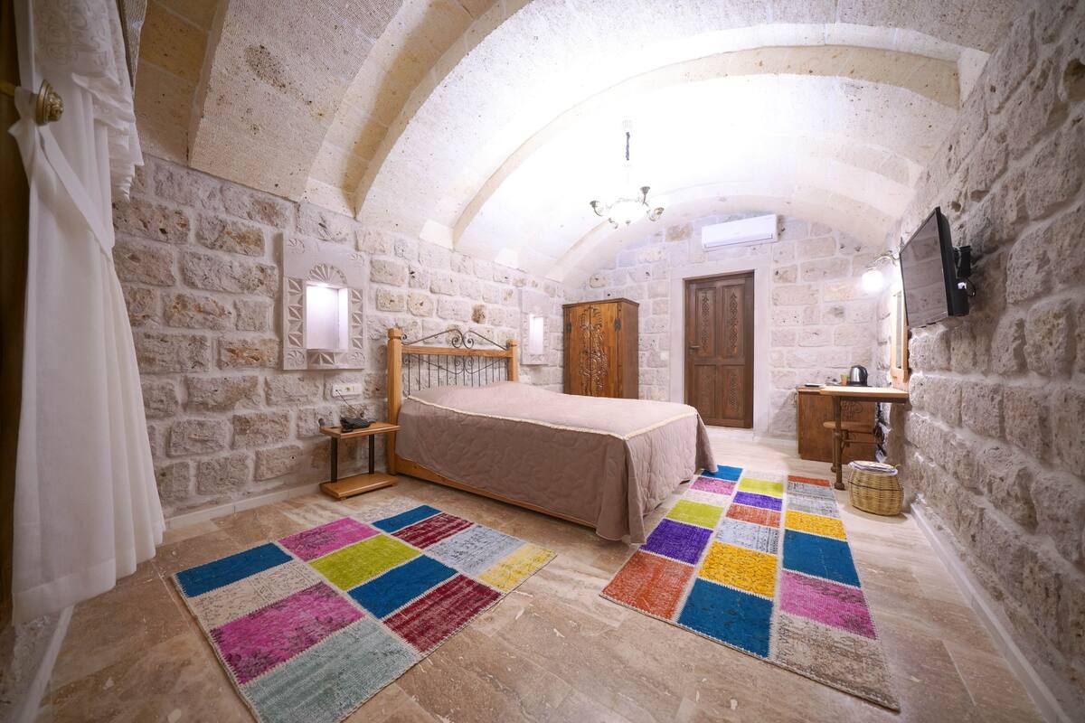 A bedroom with a stone wall, a wooden bed, a colorful rug, and a small wooden table.