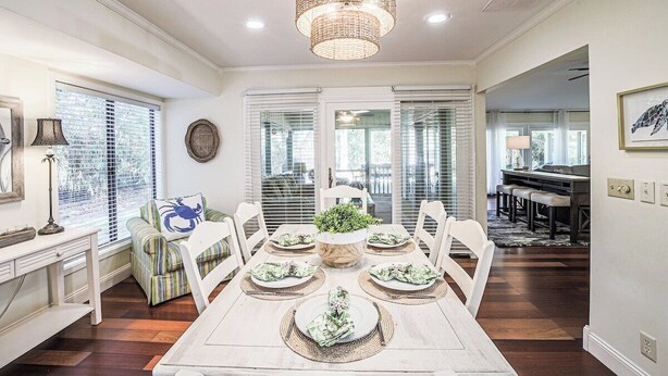 A dining area with a white table set for six, surrounded by white chairs, a woven chandelier, and a potted plant in the center.