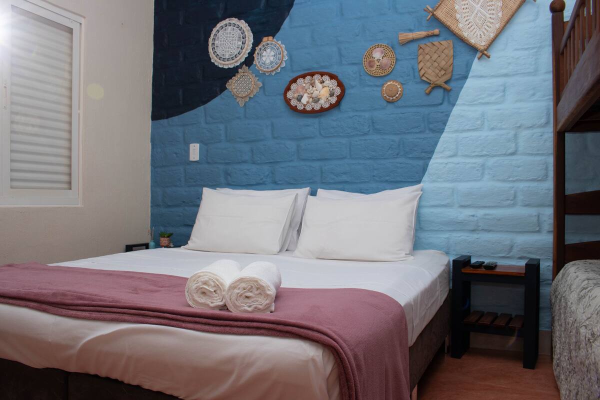A neatly made bed with white linens and a pink blanket, a bedside table with a remote control, and a wall decorated with woven wall hangings.