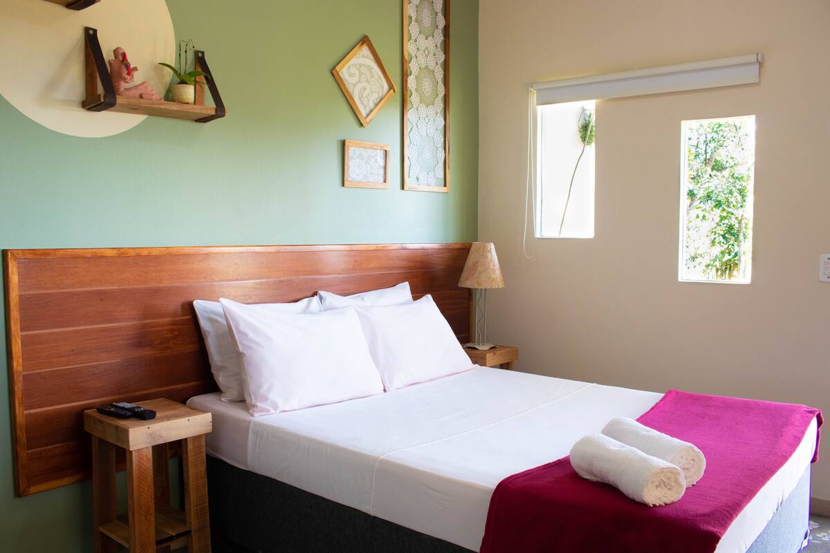 A neatly made bed with white linens, a wooden headboard, and a red throw blanket. There is a small wooden nightstand with a lamp and a remote control on it. The room has light green walls and a window with a view of greenery outside.