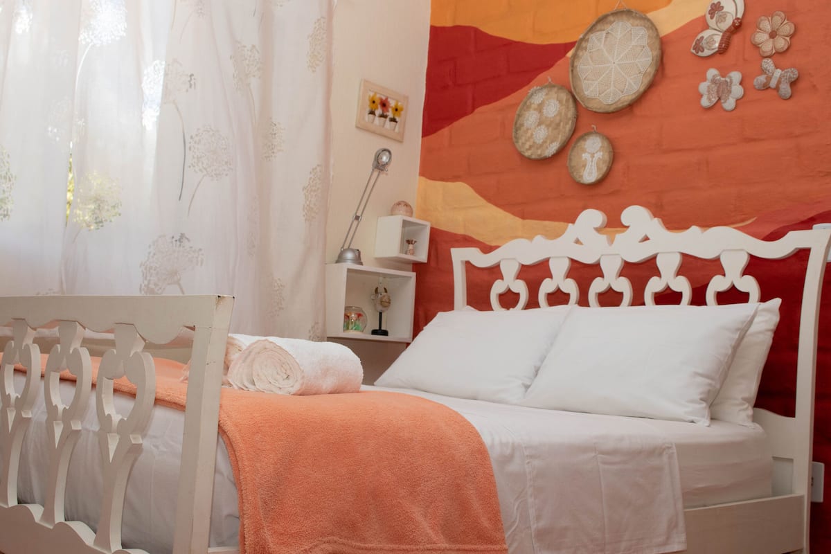 A neatly made bed with white bedding and an orange blanket, a white headboard, and a wall with decorative plates and a shelf with a lamp and small items.