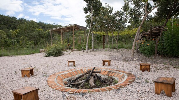 Un hogar circular rodeado de taburetes de madera, ubicado en una zona de grava con un pérgola de madera y árboles al fondo.