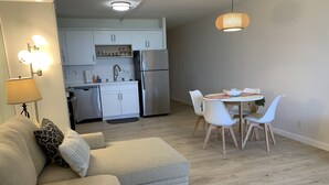 Ein modernes Wohnzimmer mit einer Couch, einem Esstisch mit Stühlen, einer Küchenzeile mit Schränken und Geräten sowie einer Pendelleuchte.