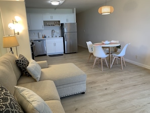 A modern living room with a sofa, a dining table with chairs, a kitchen area with cabinets and appliances, and a hanging light fixture.