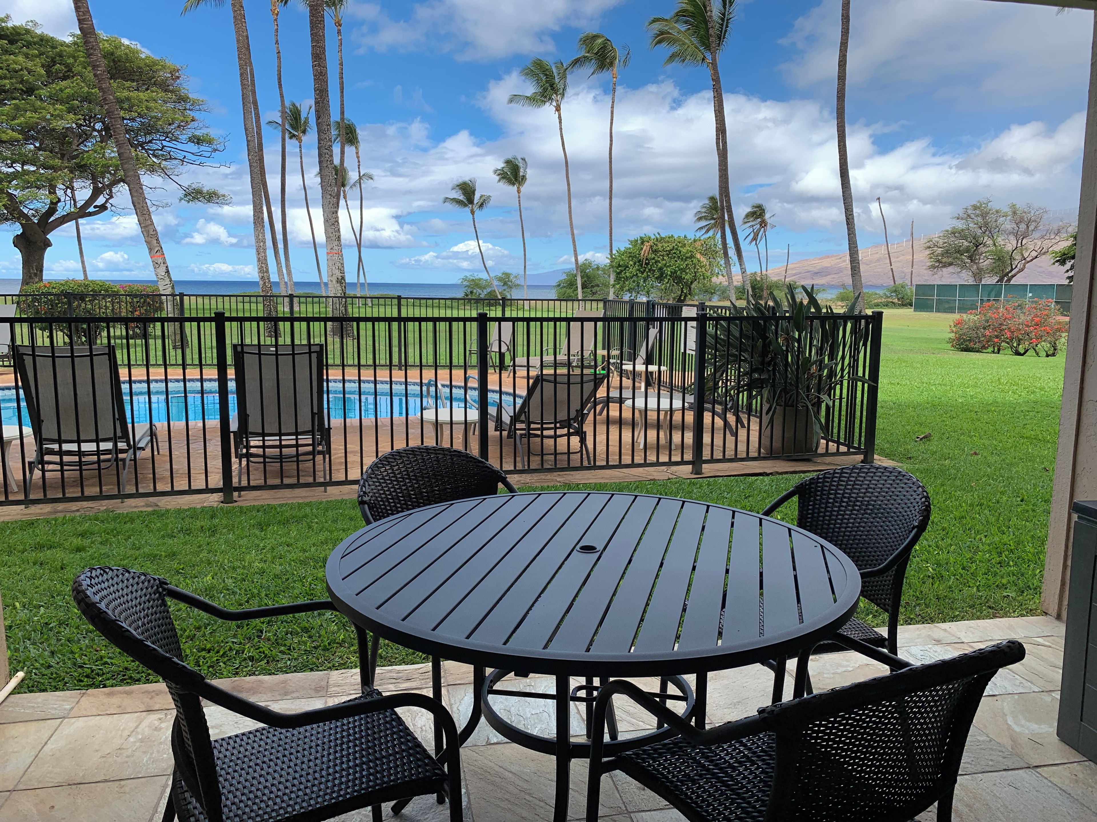 A patio with a round table and chairs, a swimming pool, and palm trees.