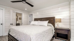 A bedroom with a wooden headboard, a bed with white linens, a nightstand with a lamp, and a ceiling fan. - Stunning Ocean Front Condo (Oceans 6) (Pine Knoll Shores)