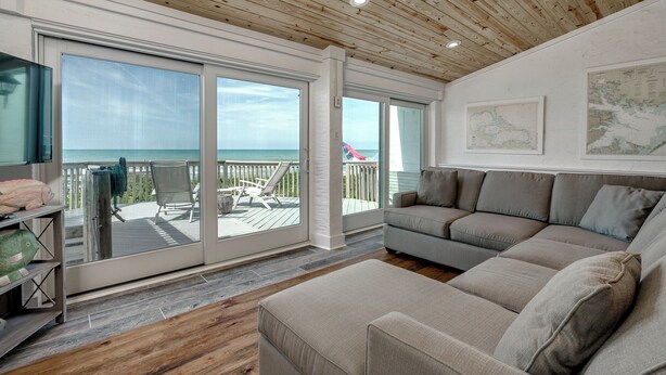 A spacious living room with a large window overlooking the ocean, a wooden ceiling, and a cozy seating area. - Stunning Ocean Front Condo (Oceans 6) (Pine Knoll Shores)