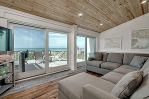 A spacious living room with a large window overlooking the ocean, a wooden ceiling, and a cozy seating area. - Stunning Ocean Front Condo (Oceans 6) (Pine Knoll Shores)