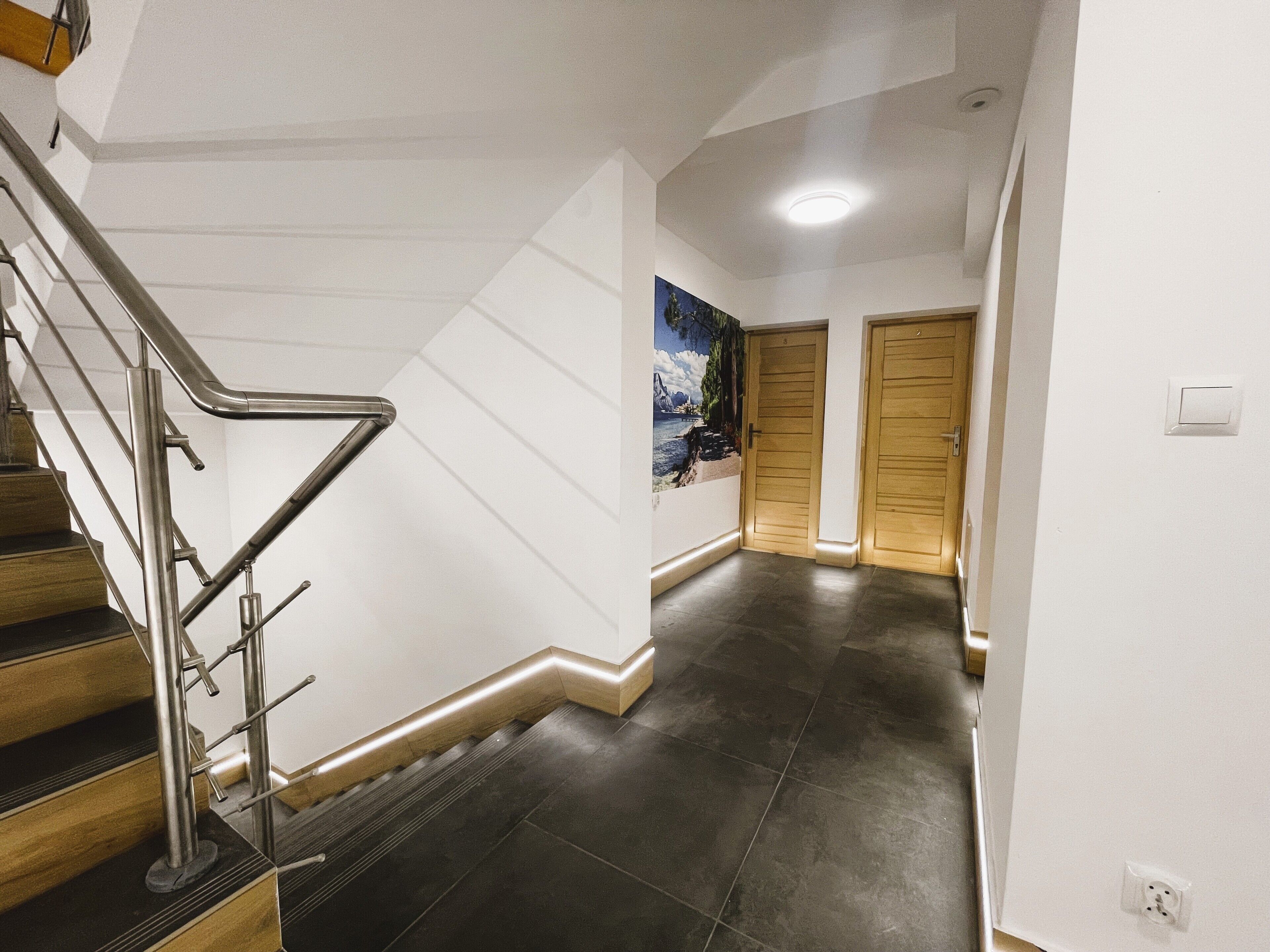 A modern interior with a wooden staircase, dark tiled flooring, and a hallway leading to two wooden doors.