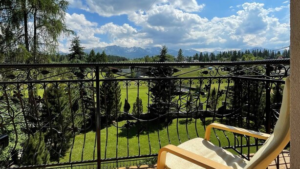 A wooden deck with a metal railing, a wooden chair, and a view of a green field and mountains in the distance. - Pokoje Gościnne Magnas (Poronin)