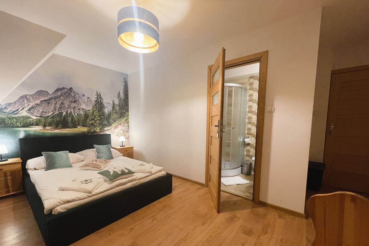 A bedroom with a bed, a nightstand, a lamp, a wooden chair, a wooden door, and a shower area.