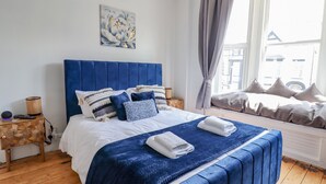 A bedroom with a large bed, a headboard, a bedside table, a lamp, and a window with curtains.