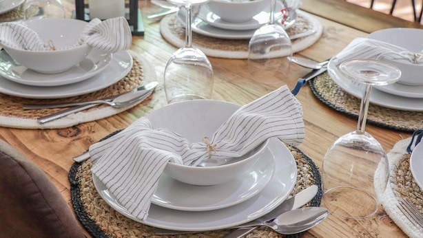 A dining table set with white plates, bowls, and cutlery, with woven placemats and striped napkins.
