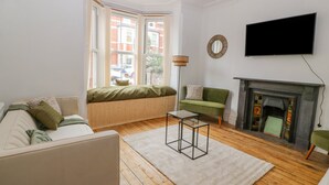 A living room with a sofa, a green armchair, a glass coffee table, a fireplace, and a TV mounted on the wall.