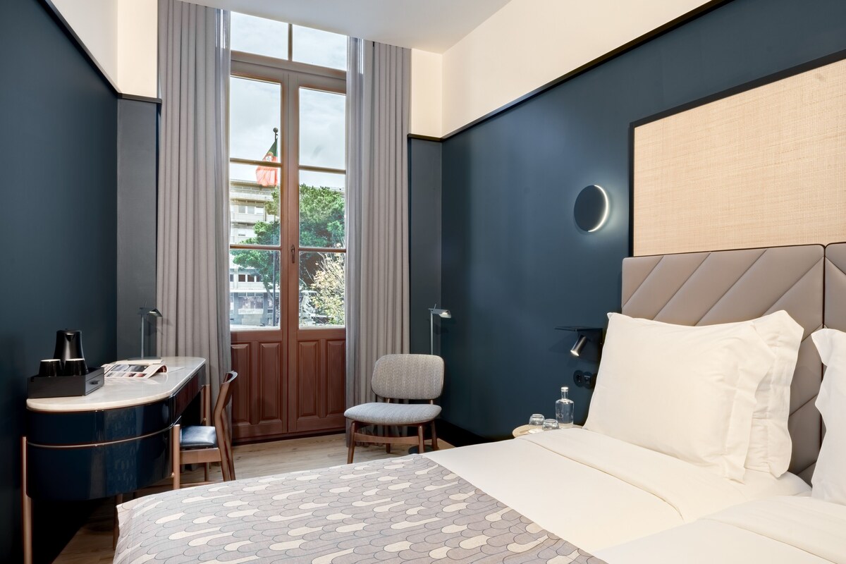 Moderni hotellihuone, jossa on suuri sänky, työpöytä ja tuoli. Huoneessa on ikkuna, jossa on verhot, ja ikkunasta avautuu näkymä ulos. Seinällä on pyöreä valaisin.