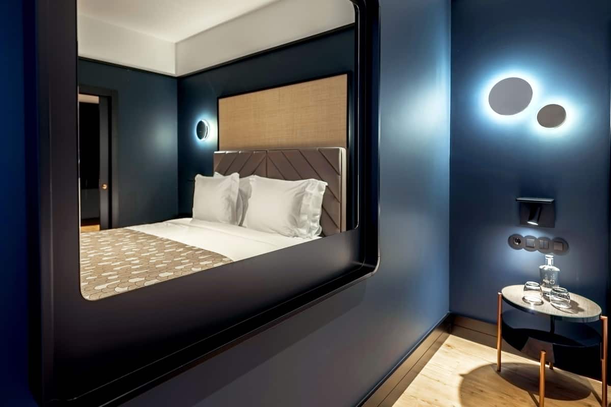A modern bedroom with a large bed, a round mirror, a small table with glasses, and a wall-mounted light fixture.