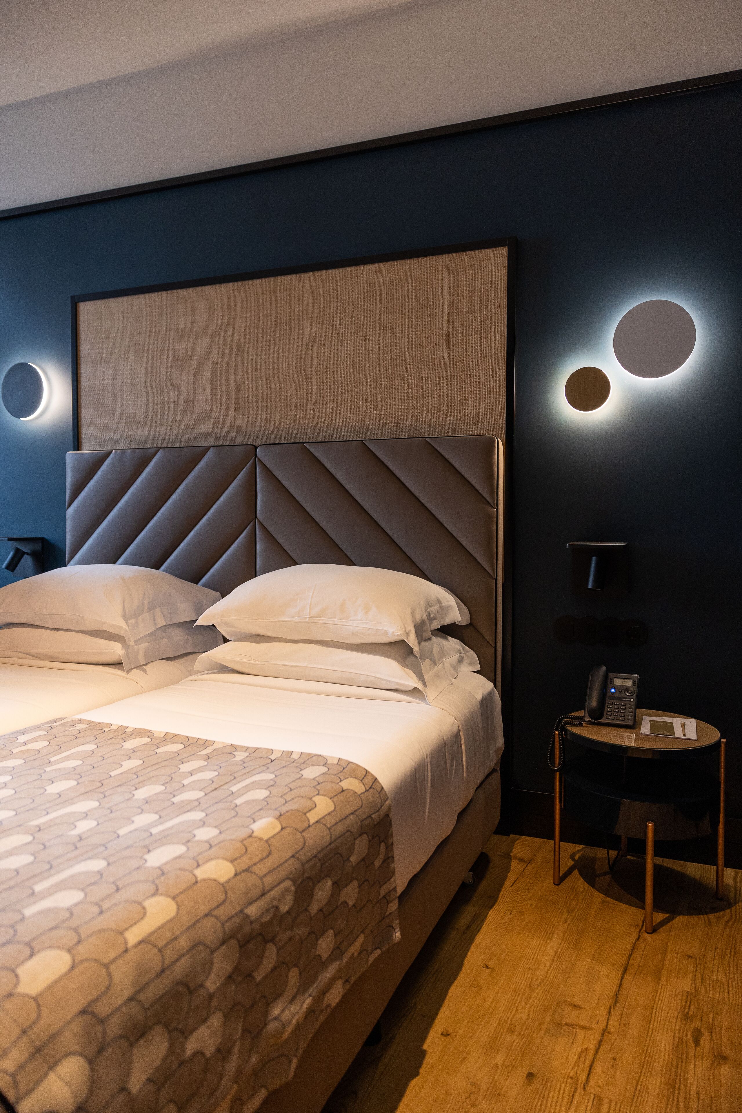A modern hotel room with a large bed, a headboard with a geometric pattern, a bedside table with a phone and a lamp, and a wall-mounted light fixture.