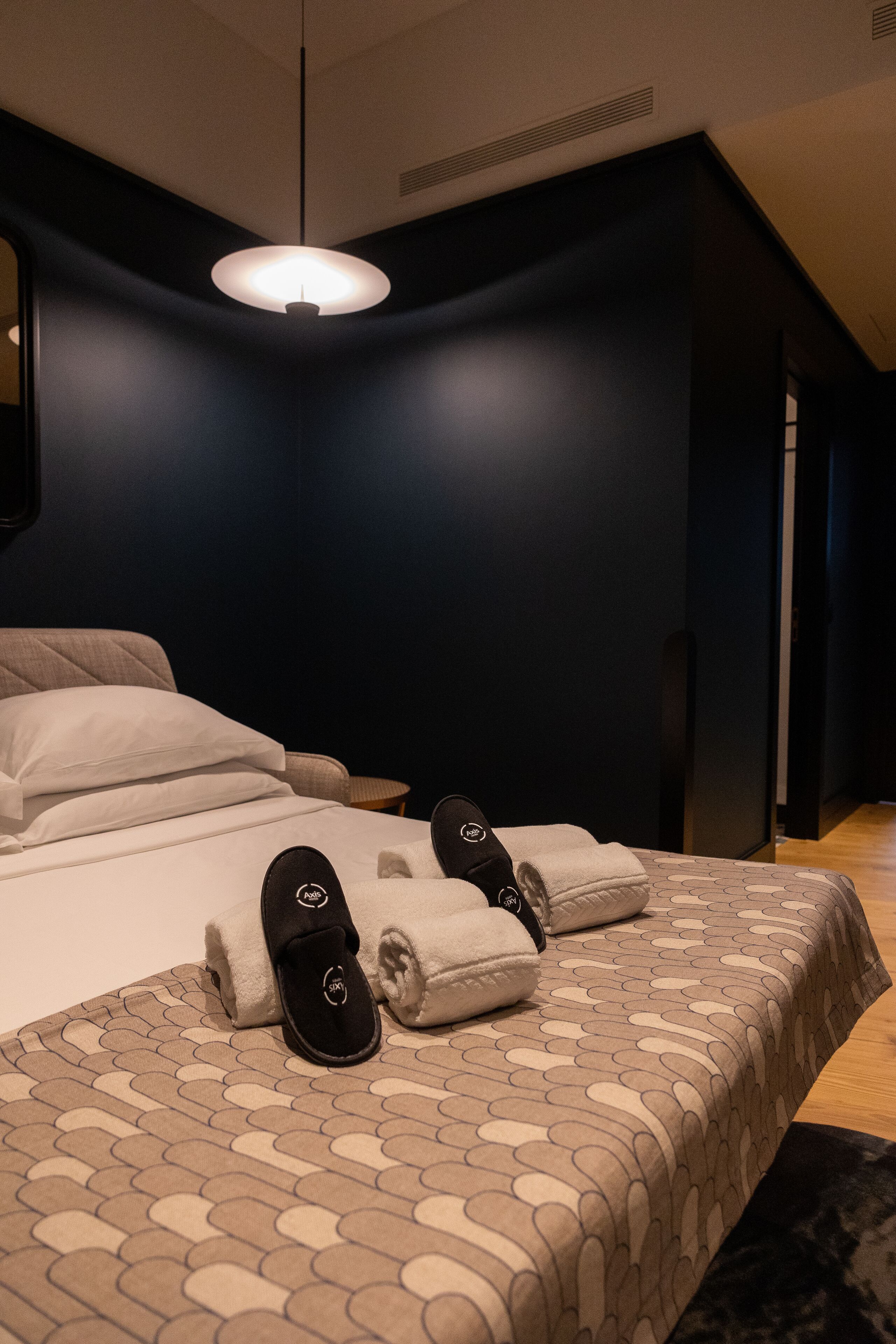 A modern hotel room with a neatly made bed, a hanging light fixture, and a dark wall.