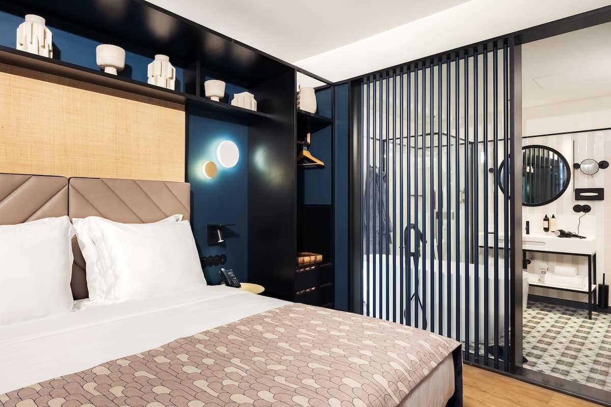 A modern hotel room with a large bed, a wardrobe with hanging clothes, a bathroom with a mirror and sink, and a sliding door leading to a shower area.