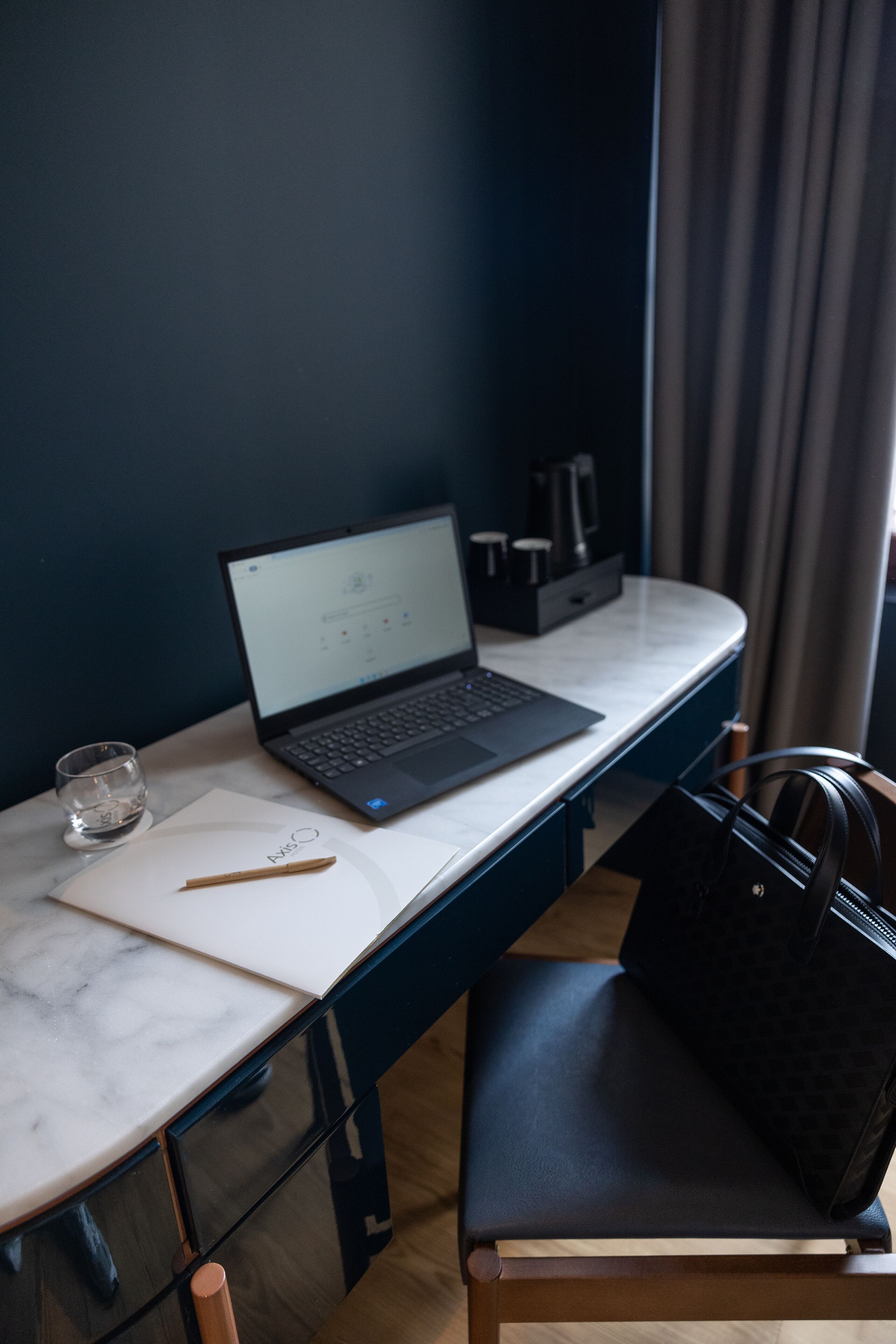 A laptop on a marble desk with a glass of water, a pen, and a notebook. A black handbag is on a chair beside the desk.