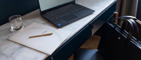 A laptop on a marble desk with a glass of water, a pen, and a notebook. A black handbag is on a chair beside the desk.