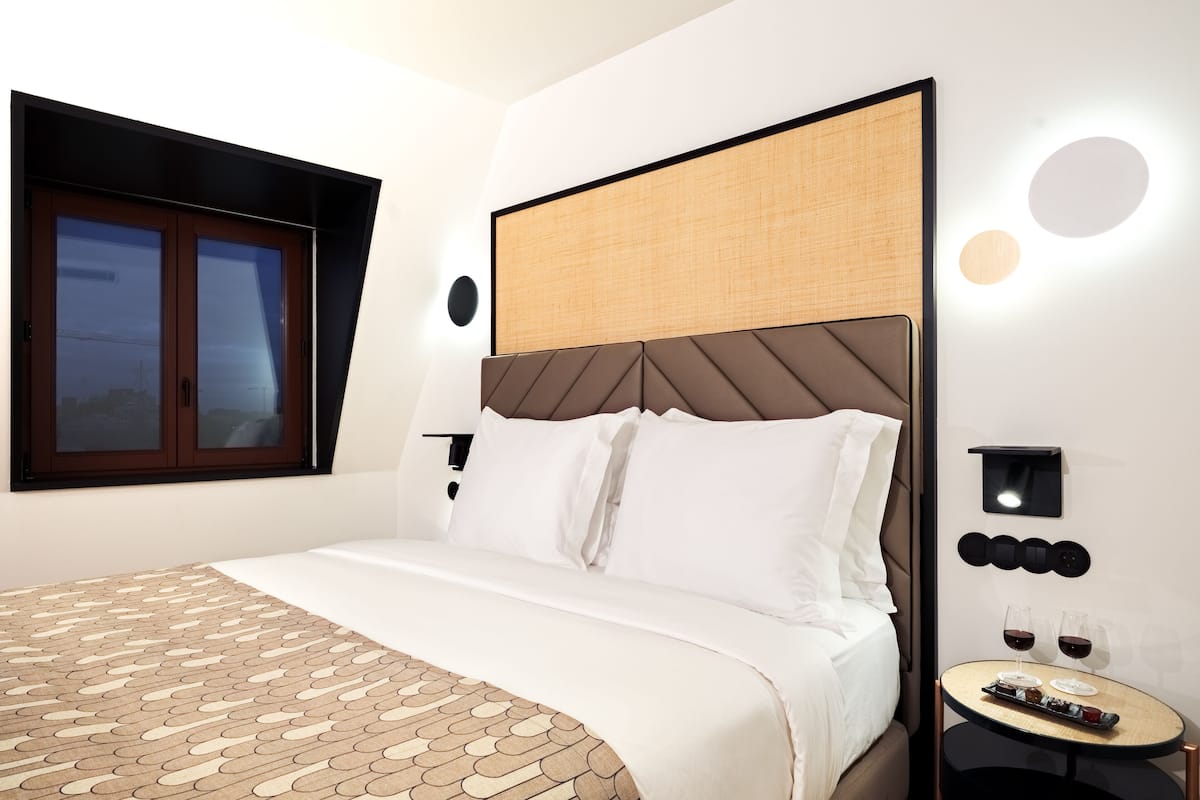 A modern hotel room with a large bed, a small side table with glasses of wine, and a window with a view of the city.