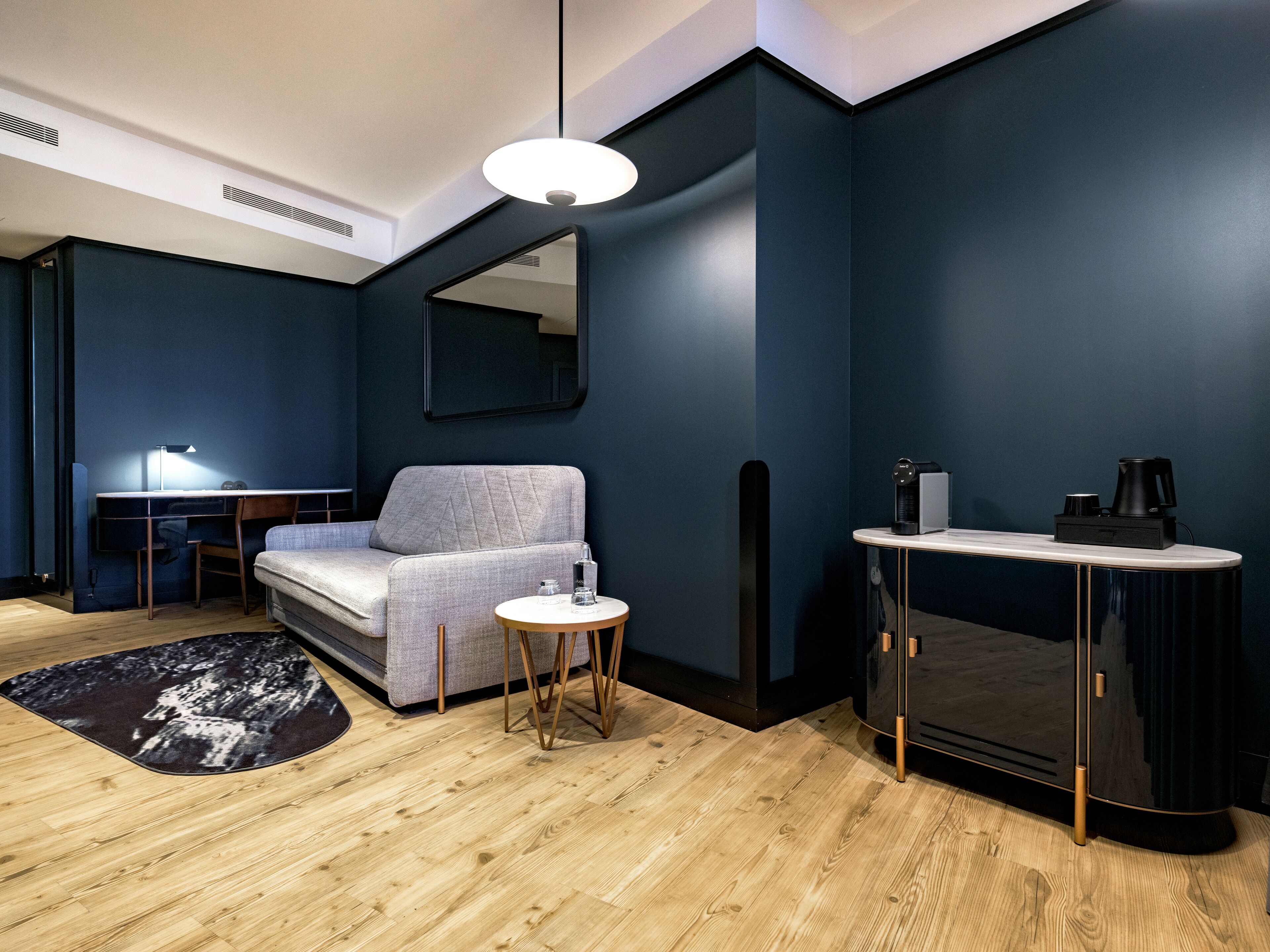 A modern room with a dark blue wall, a grey sofa, a wooden floor, a small round table, and a black cabinet with a coffee machine.