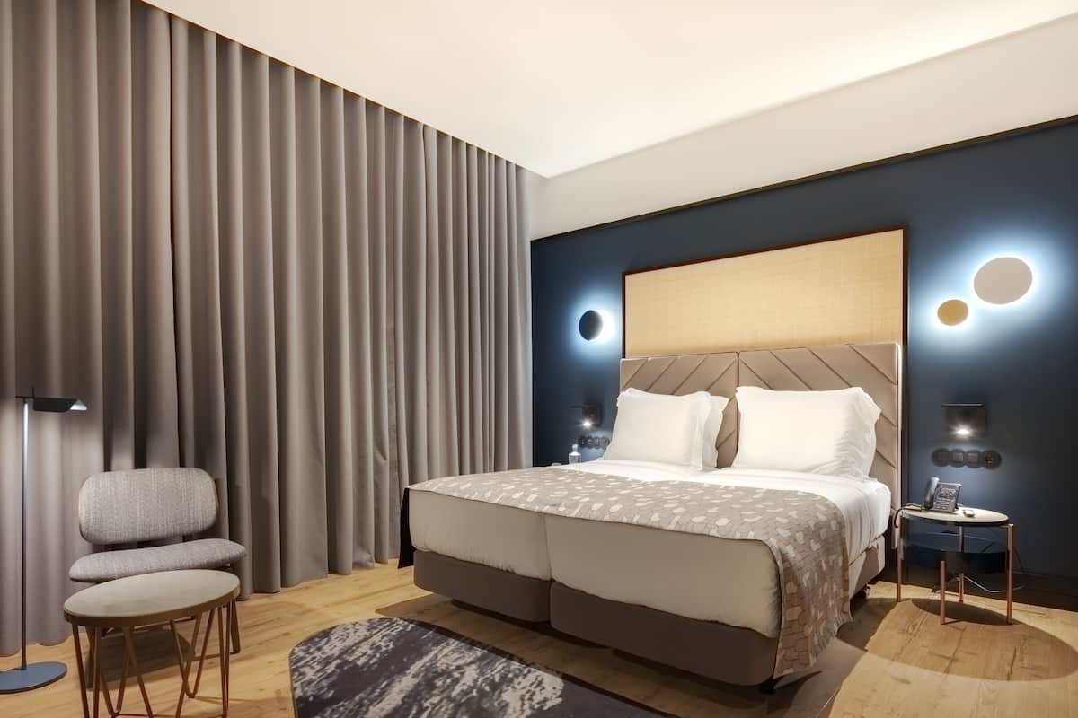 A modern hotel room with a large bed, a seating area with a chair and table, and a bedside table with a lamp and phone.