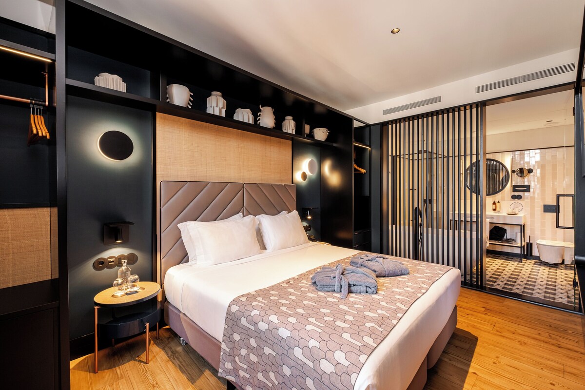 Moderni hotellihuone, jossa on suuri sänky, yöpöytä lampun kanssa ja peilillä varustettu vaatekaappi.
