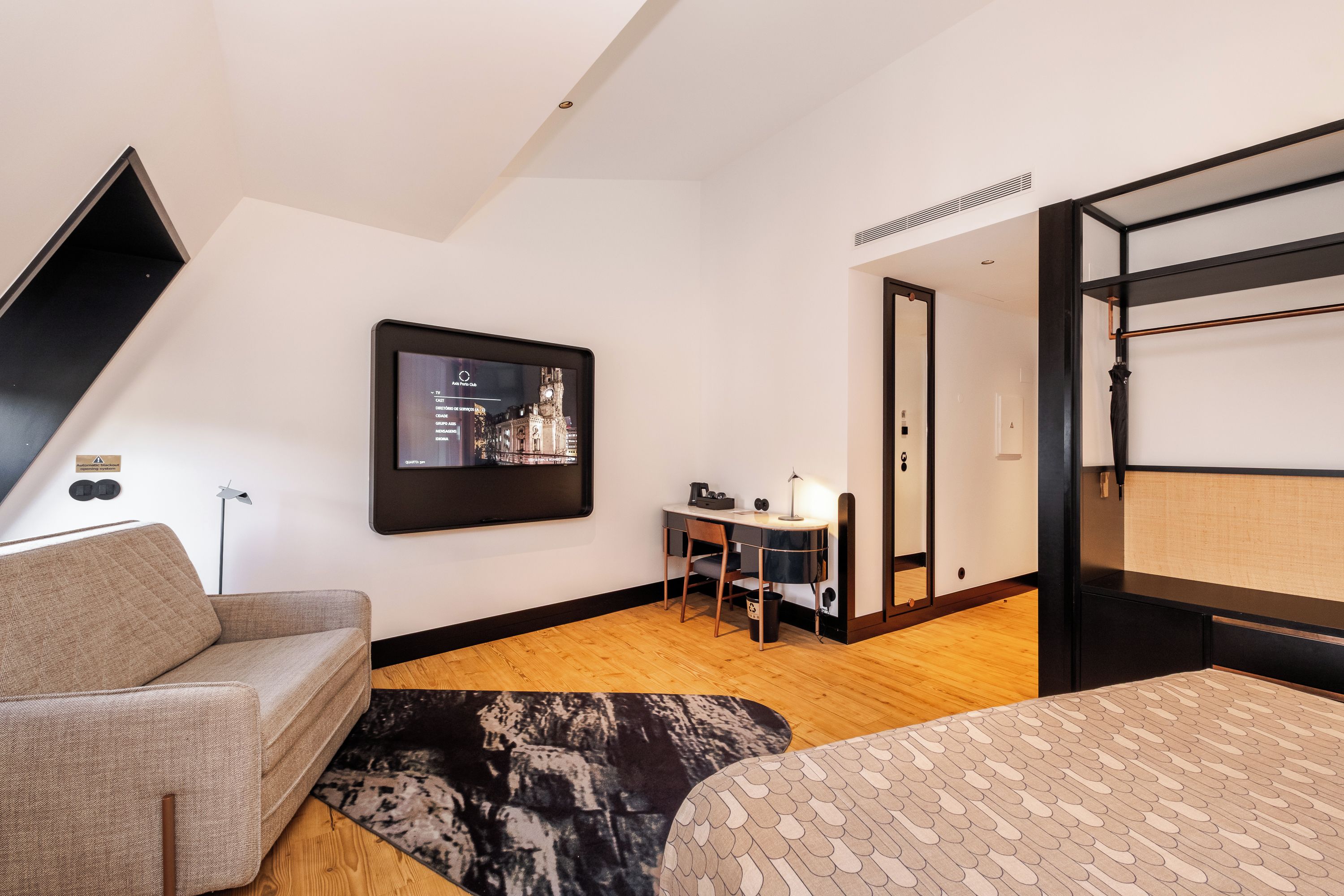 A modern hotel room with a bed, a desk, a chair, a TV mounted on the wall, and a view of the cityscape.