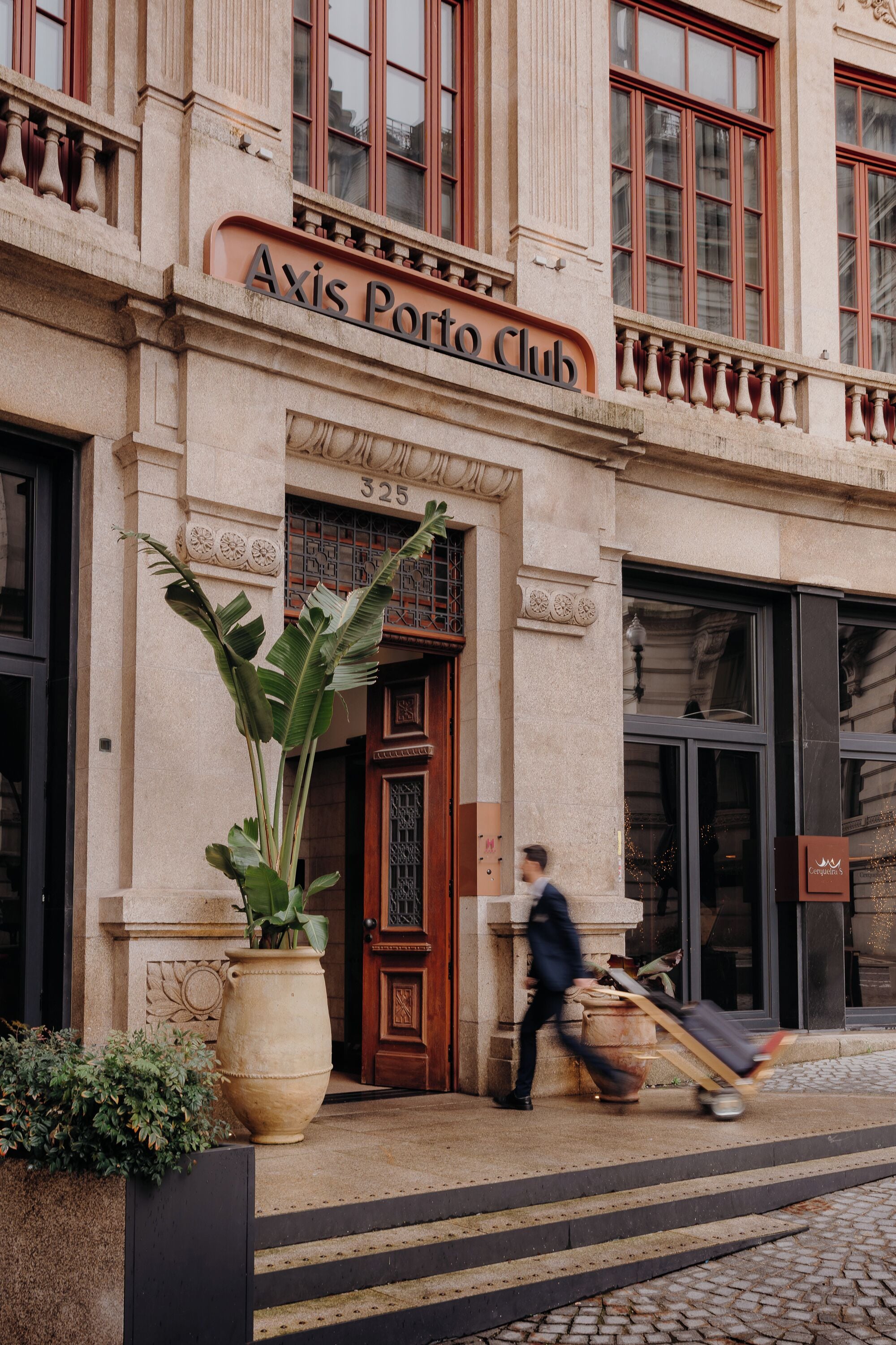 The exterior of the Axis Porto Club, featuring a large planter, a wooden door, and a person pushing a cart.