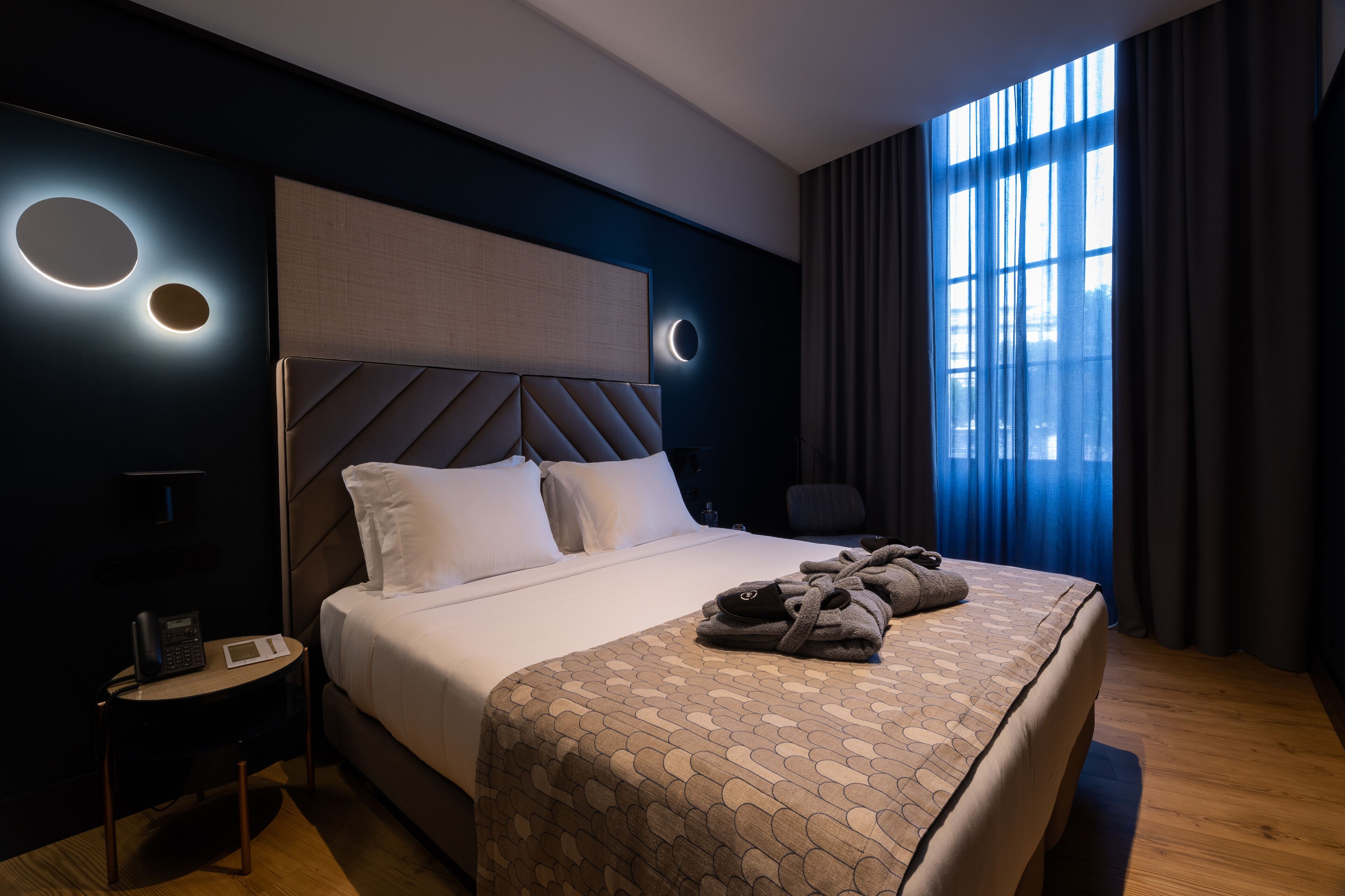 A modern hotel room with a large bed, a bedside table with a phone and a lamp, and a view of the city through large windows.
