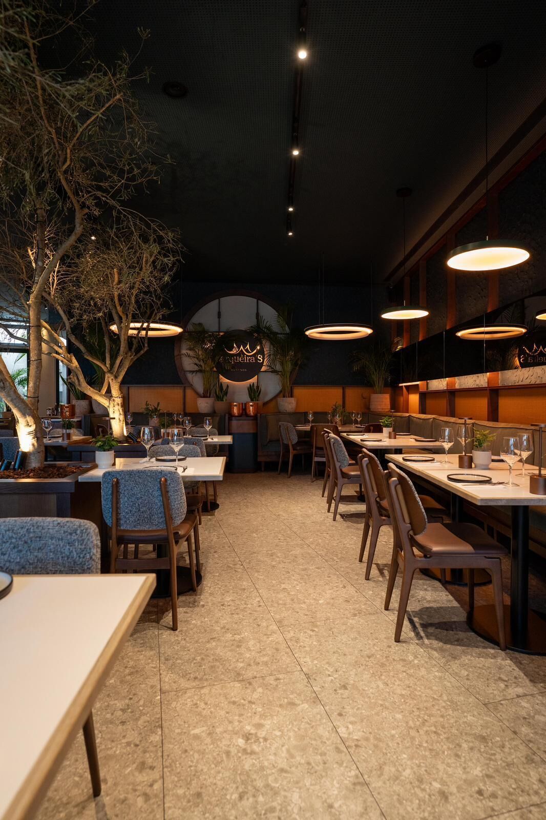 A restaurant with a central bar area, multiple dining tables, and chairs. There are hanging lights and a tree in the center.