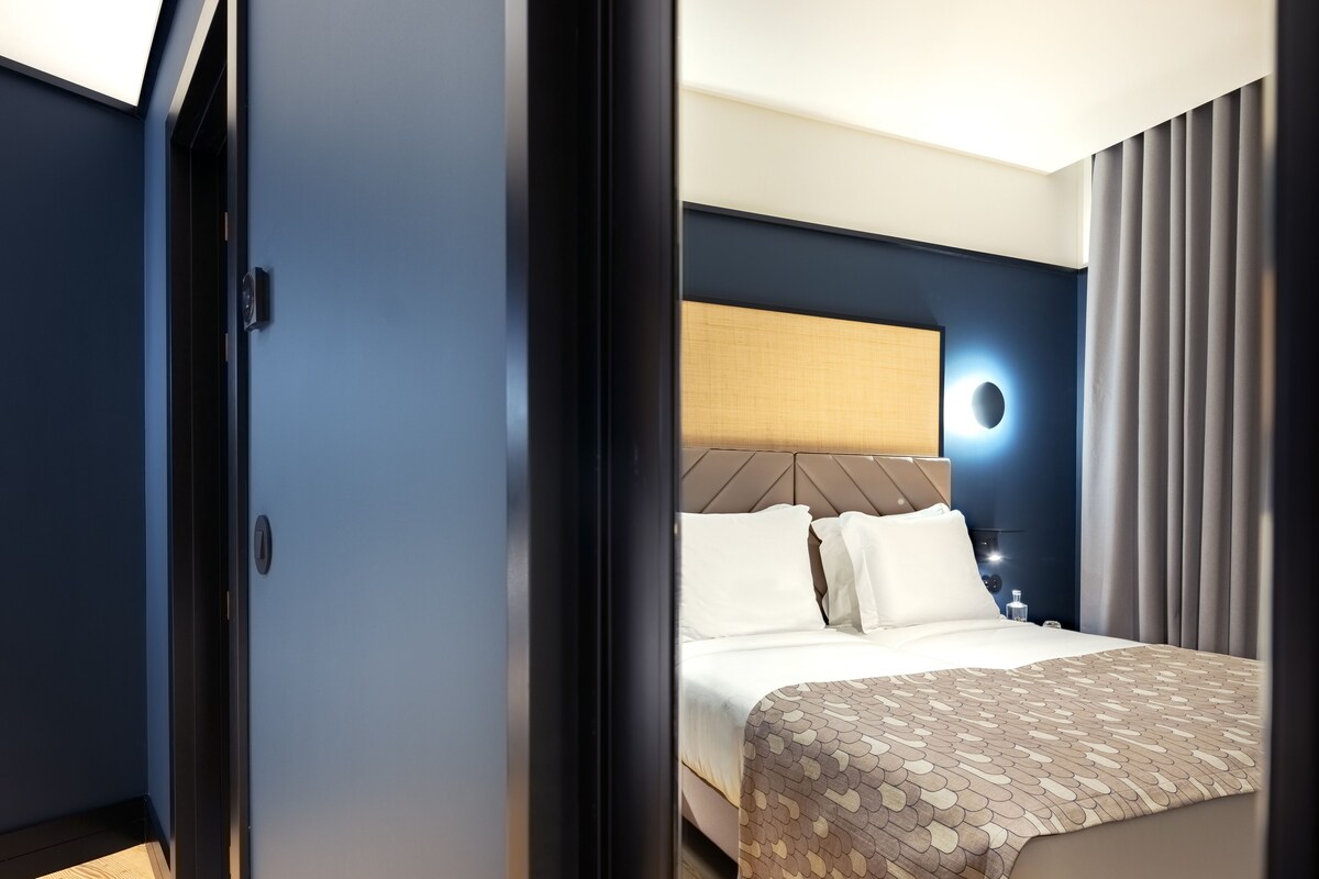 Hotellihuone, jossa on sänky, sängynpääty, yöpöytä, lamppu ja ikkuna, jossa on verhot.