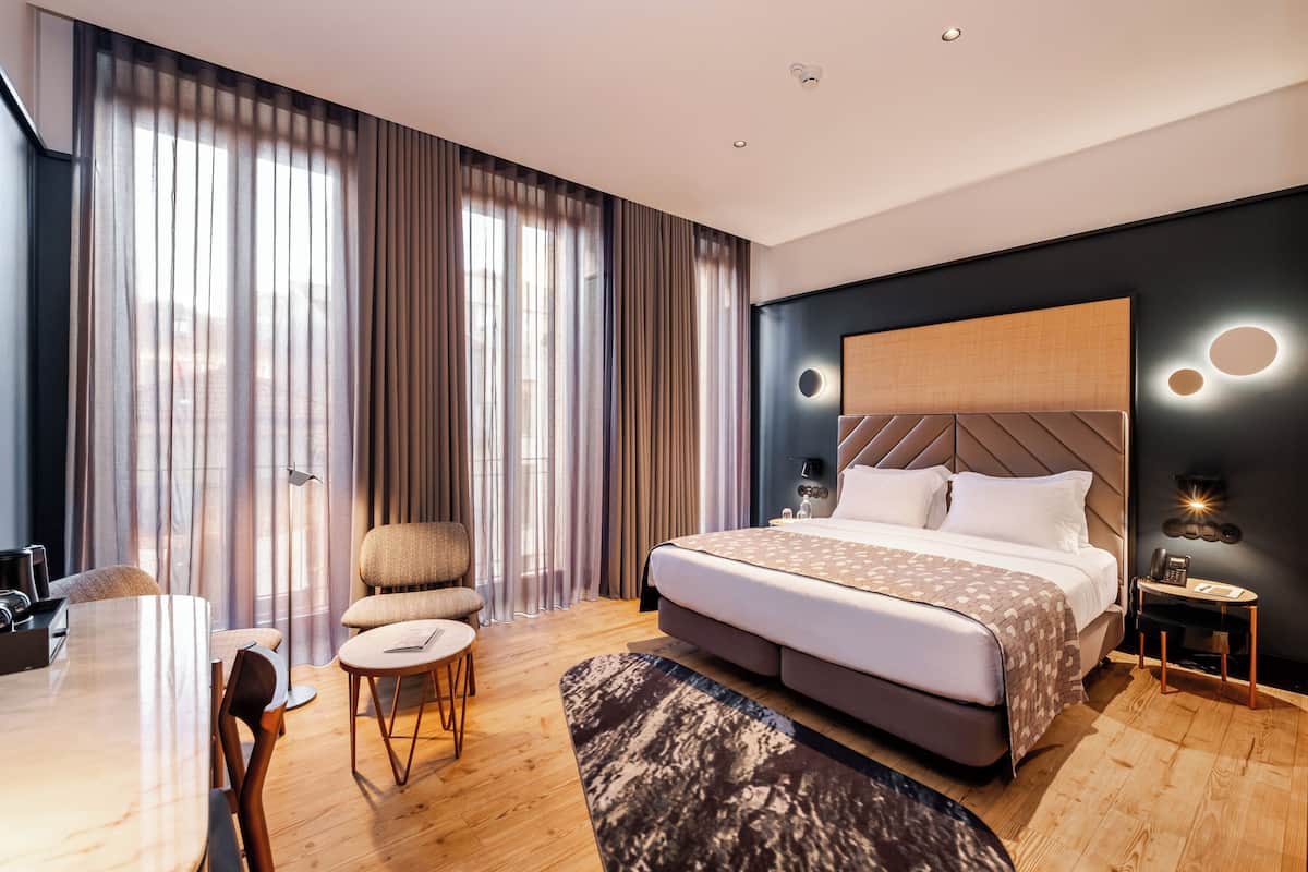 A modern hotel room with a large bed, a desk, a chair, and a view of the city through sheer curtains.