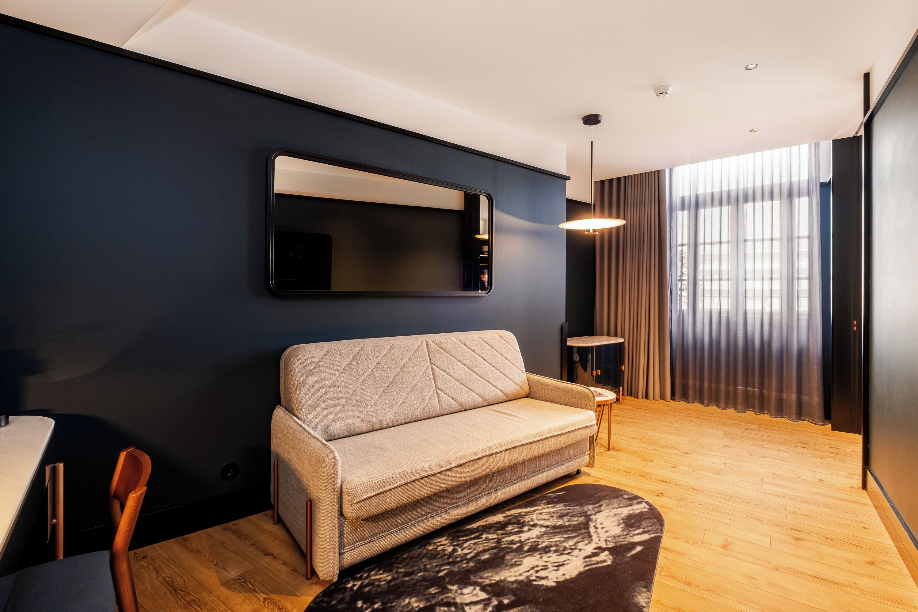 A modern living room with a sofa, a small side table, a mirror, and a window with curtains.
