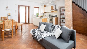 Una sala moderna con un sofá gris, una mesa de comedor de madera y sillas. Hay una zona de cocina con armarios blancos y una pared de ladrillo.
