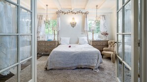 Une chambre à coucher avec un grand lit, un baldaquin, un lustre et une vue sur le paysage par la fenêtre ouverte.
