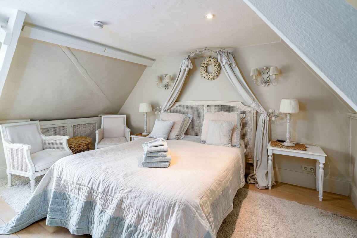 Een slaapkamer met een groot bed, een witte stoel, een nachtkastje en een wandlamp.