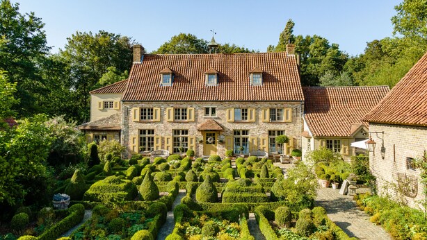 Un grand bâtiment en pierre, surmonté d’un toit de tuiles, entouré de jardins bien entretenus et bordé d’un chemin pavé.