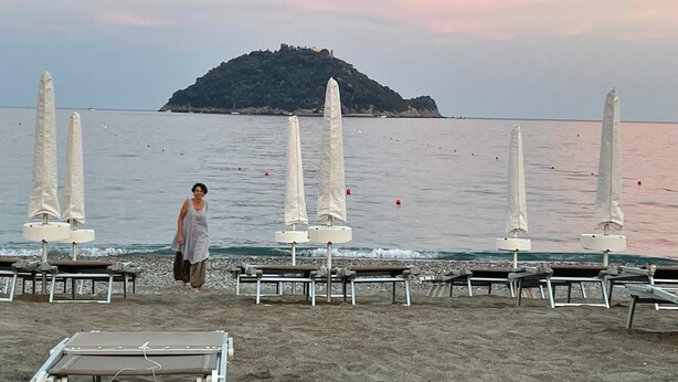 A beach with rows of sun loungers and umbrellas, a person walking, and a small island in the background.