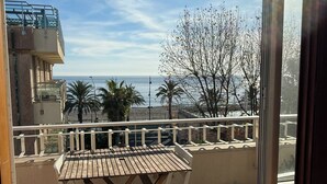 A balcony with a wooden table and chairs, overlooking a coastal view with palm trees and a clear sky.