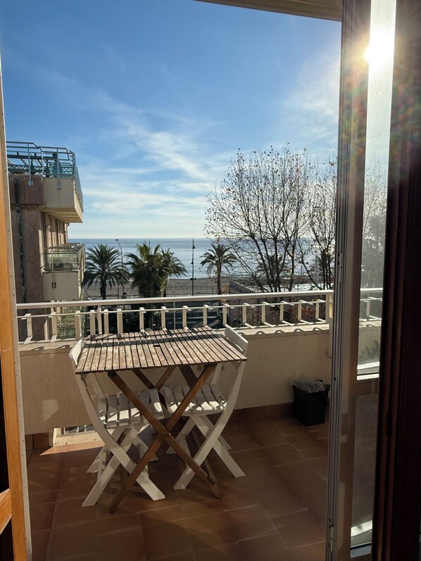 Un balcón con una mesa y sillas de madera, con vista al mar, palmeras y un cielo despejado.